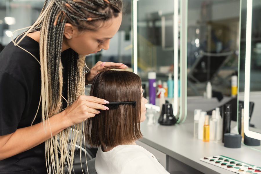 Stylist with long braids combing a client's glossy brown bob at a lit mirror