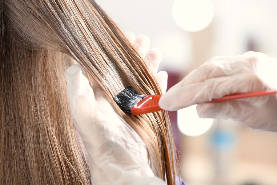Colorist in gloves brushing color onto a section of long, light-brown hair