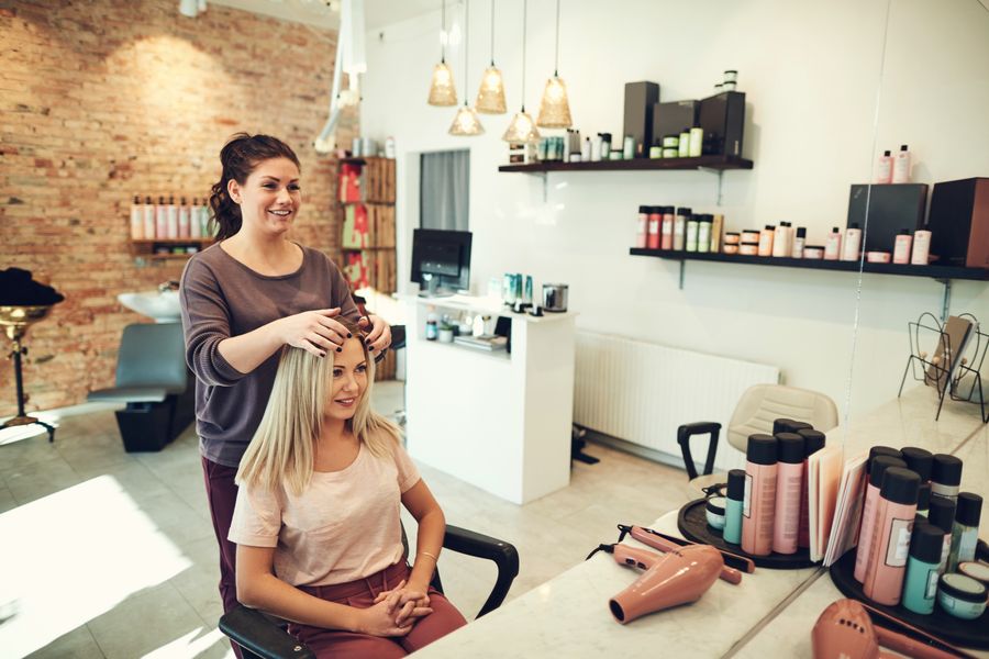 Stylist talking with a blonde client in a sunny salon with brick walls, pendant lights and product shelves