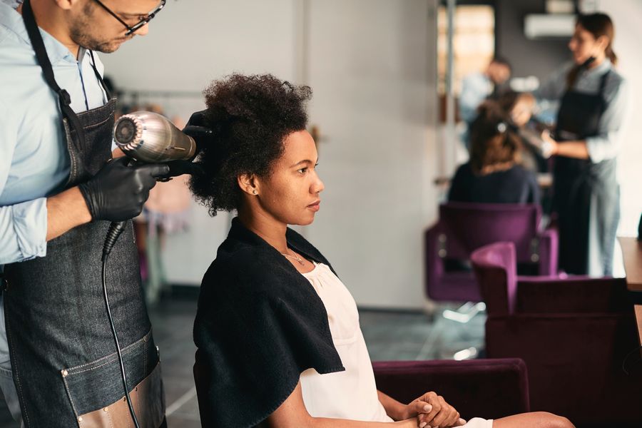 Stylist blow-drying a client's natural hair in a bright, busy salon