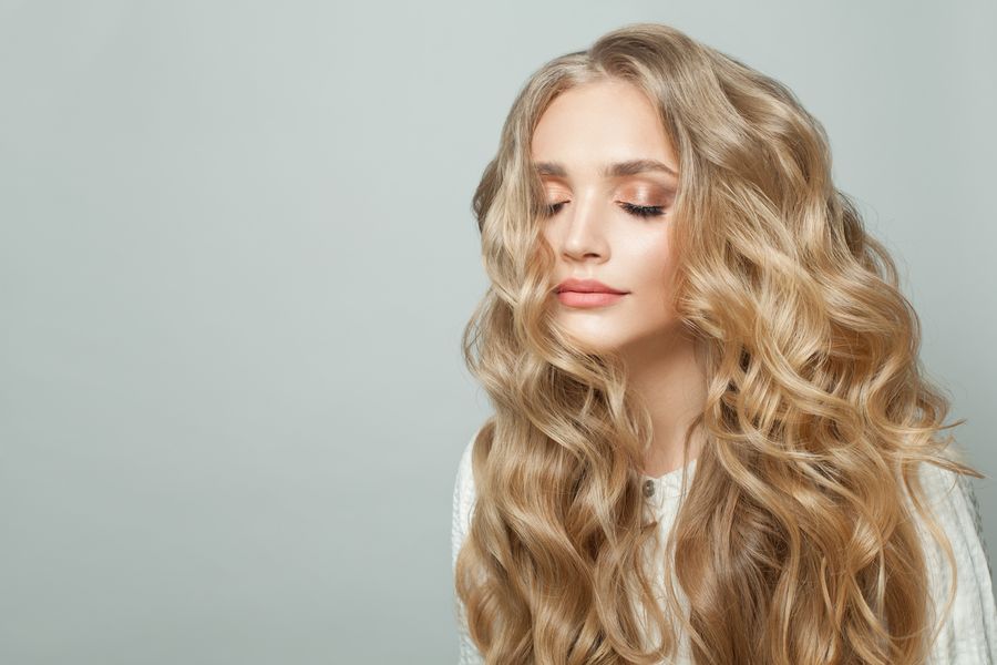 Woman with long, voluminous golden blonde curls, eyes closed, against a pale gray-green background