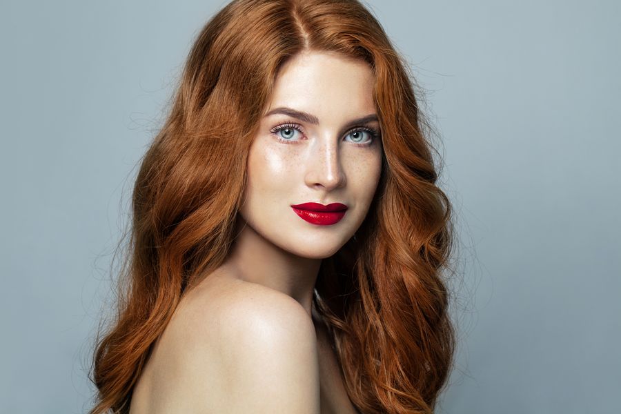 Woman with long copper-red waves, freckles and red lipstick against a soft blue-gray background