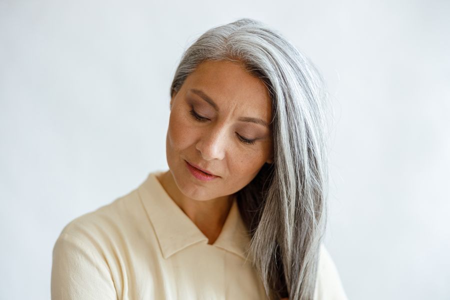 Middle-aged Asian woman with long, sleek silver hair in a cream blouse, looking down serenely